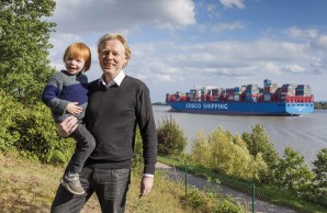 Prof. Dr. Jürgen Gallinat steht am oberen Elbufer, auf dem Arm trägt er seinen kleinen, rothaarigen Sohn, beide lächeln in die Kamera. Im Hintergrund die Elbe, blauer Himmel mit wenigen Wolken, ein voll beladenes Containerschiff fährt Richtung Hamburger Hafen