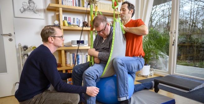 Tomas Gerlach in einem Tuch, das ihn im Sitzen aufrecht hält, dabei sein Assistent und sein Physioterapeut